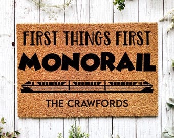 May include: A rectangular doormat made of natural coir fiber. The mat features the text "FIRST THINGS FIRST MONORAIL" in bold black letters, with a monorail graphic below. The family name "THE CRAWFORDS" is printed at the bottom.