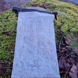 Könnte beinhalten: Eine hellgraue rechteckige Tasche mit schwarzem Riemenverschluss. Die Tasche besteht aus einem strukturierten Material und trägt den Aufdruck "STARK NORDIK MADE IN SWEDEN". Die Tasche liegt auf einer moosigen Oberfläche.