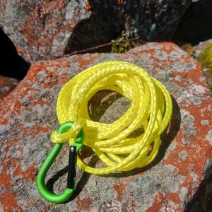 May include: A coiled yellow rope with a green carabiner clip. The rope is looped and knotted, and the carabiner is attached to the end. The items are resting on a reddish-brown rock.