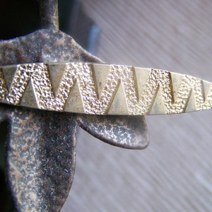 May include: A gold-toned metal bar with a textured surface and a repeating geometric pattern. The bar has a slightly curved shape.