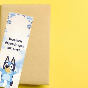 May include: A bookmark with the text "Happiness depends upon ourselves" and a cartoon character illustration. The bookmark has a blue and white floral design at the top. It rests on a brown rectangular object, set against a bright yellow background.