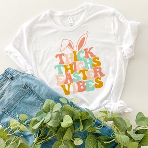 May include: White t-shirt with a graphic design of a bunny with the text "Thick Thighs Easter Vibes" in colorful retro font.