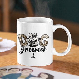 May include: White ceramic mug with a black and brown paw print design. The text "Dog groomer" is written below the paw print. A pair of scissors is below the text.
