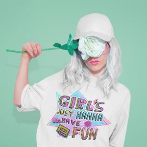 May include: A white sweatshirt with a pink, blue, and yellow graphic print that says "Girls Just Wanna 80s Have Fun" with a cassette tape graphic. The model is wearing a white baseball cap and has white hair.