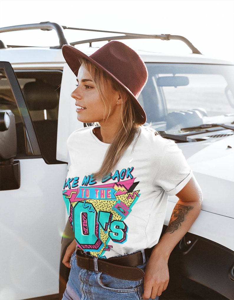 May include: A white t-shirt with a retro 80s graphic design featuring the text "Take Me Back To The 80s" in a colorful, geometric style. The design includes a cassette tape and a pink and blue color scheme.