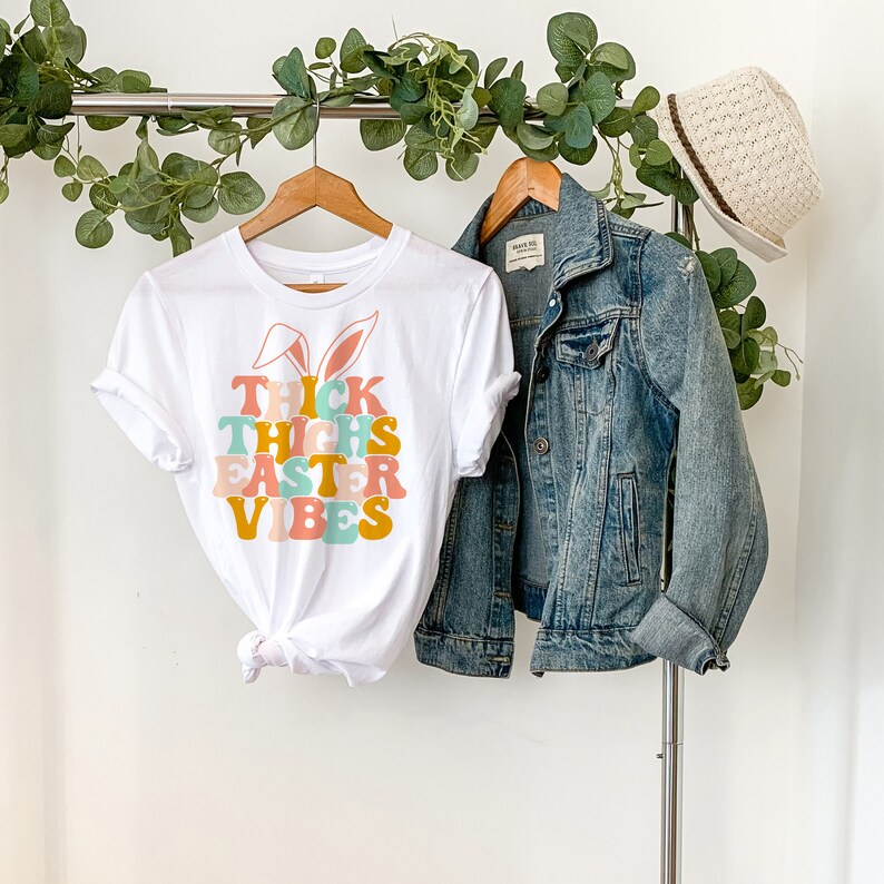 May include: White t-shirt with a graphic of bunny ears and the text "Thick Thighs Easter Vibes" in colorful retro font.
