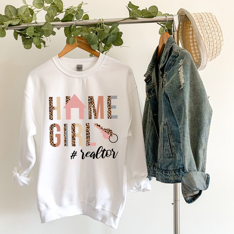 K&ouml;nnte beinhalten: Ein wei&szlig;es Sweatshirt mit Rundhalsausschnitt und dem Schriftzug HOME GIRL, bei dem ein Haussymbol das O und ein Schl&uuml;sselsymbol das I ersetzt. Die Buchstaben haben ein Leopardenmuster und der Text #realtor steht in schwarzer Schreibschrift darunter.