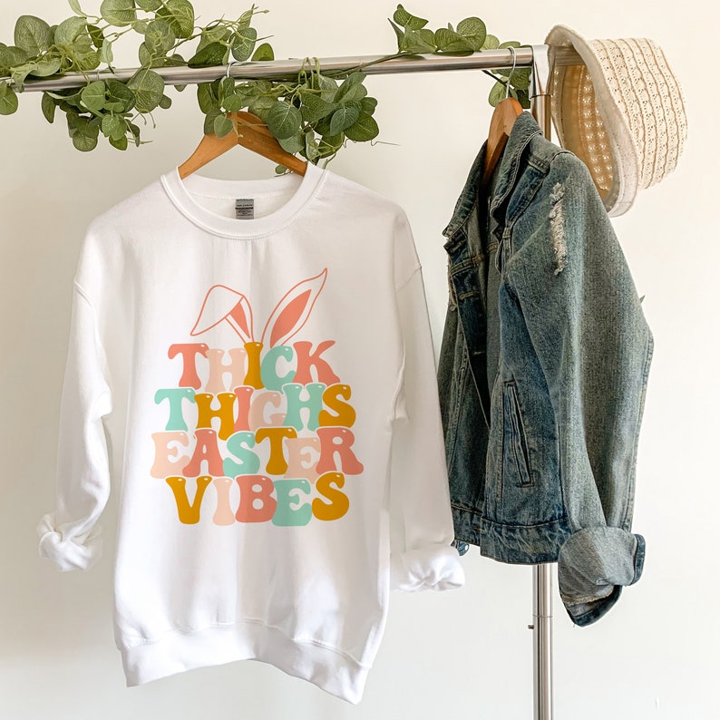 May include: White sweatshirt with a graphic design of a bunny with the text "Thick Thighs Easter Vibes" in colorful retro font.