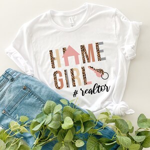 K&ouml;nnte beinhalten: Ein wei&szlig;es Kurzarm-T-Shirt mit dem Schriftzug HOME GIRL, bei dem ein Haussymbol das O und ein Schl&uuml;sselsymbol das I ersetzt. Die Buchstaben haben ein Leopardenmuster und ein beiges Design. Darunter steht der Text #realtor in schwarzer Schrift.