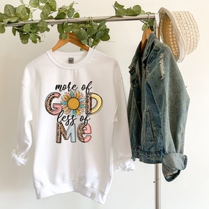 Może przedstawiać: Biała bluza z nadrukiem graficznym, kt&oacute;ry m&oacute;wi "more of God less of me" z słonecznikiem w środku. Grafika jest w kolorze r&oacute;żowym, ż&oacute;łtym, niebieskim i brązowym.