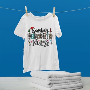 May include: White t-shirt with a Christmas-themed design that says "Santa's Favorite Nurse" in colorful letters with a Santa hat, Christmas lights, and Christmas trees.