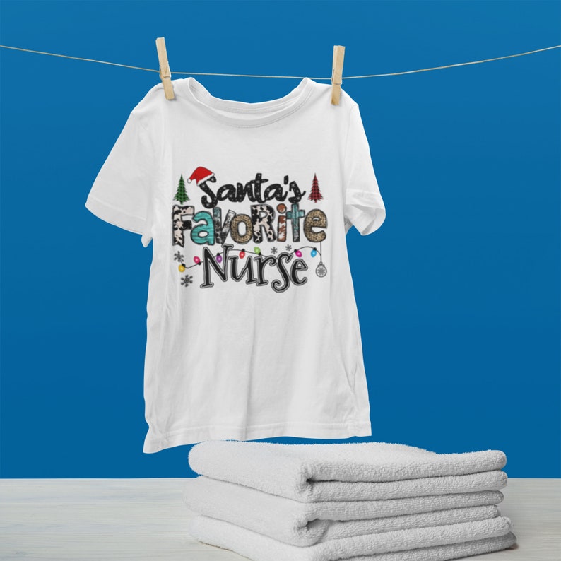 May include: White t-shirt with a Christmas-themed design that says "Santa's Favorite Nurse" in colorful letters with a Santa hat, Christmas lights, and Christmas trees.