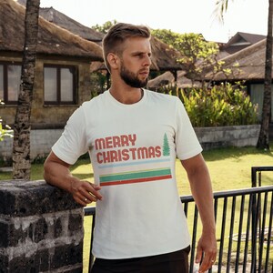 May include: A white t-shirt with a retro-style graphic design that says "Merry Christmas" with a stylized Christmas tree. The design features red, blue, green, and yellow stripes.