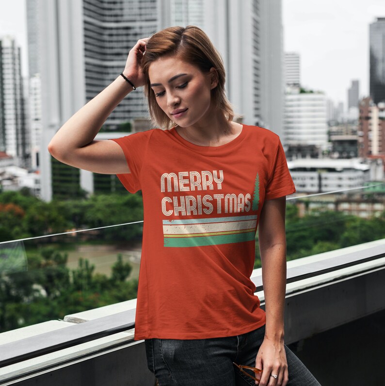 May include: A red t-shirt with a retro-style graphic that says "Merry Christmas" with a stylized Christmas tree. The graphic is in shades of green, yellow, and white.