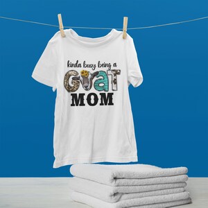 May include: White t-shirt with the text "kinda busy being a goat MOM" in black. The word "goat" is made up of a goat's head and various animal print patterns.