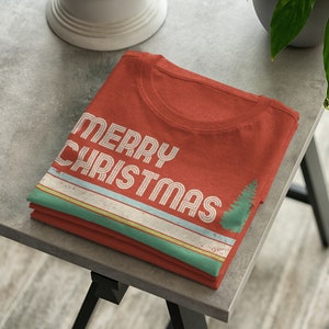 May include: A folded red t-shirt with a vintage-style "Merry Christmas" graphic and a stylized pine tree. The graphic features white, blue, and green stripes.