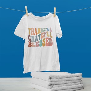 May include: White t-shirt with the words "Thankful Grateful Blessed" in a rainbow of colors. The words are in a retro font style.