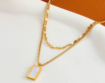 May include: A layered gold-toned necklace with a rectangular pendant. The pendant features a white, iridescent inlay. The necklace has two chains, one with a delicate link design and the other with a textured pattern. The background is a mix of white and brown.