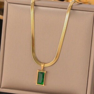 May include: Gold-coloured necklace featuring a rectangular green pendant. The pendant is an emerald green stone set in a gold frame. The chain has a flat, herringbone design. The necklace is displayed on a neutral-toned surface.