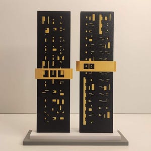 Puede incluir: Una escultura moderna con dos paneles rectangulares negros con formas recortadas y detalles dorados. Los paneles están conectados por una banda dorada con las letras "JUL". La escultura se asienta sobre una base de múltiples capas.