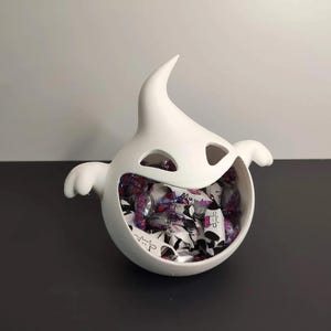 Peut inclure: Un bonbonnière blanche en forme de fantôme, avec une large bouche ouverte et des trous pour les yeux. Le plat est rempli de bonbons emballés. Le fantôme a de petits bras en forme d'ailes et un sommet pointu.