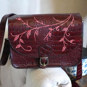 Puede incluir: Un bolso de hombro color burdeos oscuro con textura de piel de cocodrilo y cierre de solapa. La solapa frontal presenta un diseño de enredadera pintado a mano en rosa claro con hojas. Una hebilla metálica asegura la solapa y tiene correa.
