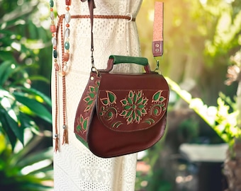 May include: A dark brown leather handbag featuring a green handle and a flap with intricate floral cutouts revealing green leather underneath. The bag includes a long adjustable shoulder strap and metal hardware, showcasing a structured, rounded silhouette.