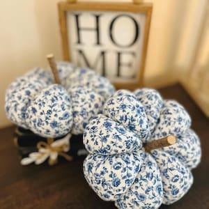 May include: Two fabric pumpkins featuring a blue and white floral pattern with a brown twig stem. Behind the pumpkins is a rectangular wooden sign with a light wood frame and a row of wooden beads on top, displaying the text HOME in black.