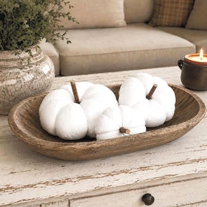 May include: An oval-shaped wooden dough bowl containing three white fabric pumpkins with brown stems. The pumpkins are arranged inside the natural wood grain bowl, which features a smooth, curved interior and a rustic, textured exterior finish.