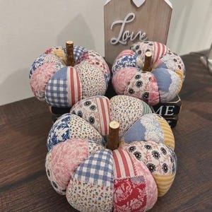 Puede incluir: Tres calabazas de tela rellenas con un diseño de patchwork de patrones florales, de cuadros vichy y de rayas en azul, rojo, rosa y amarillo. Cada calabaza tiene un pequeño tallo de madera. Detrás hay un letrero de madera en forma de casa con la palabra Love.