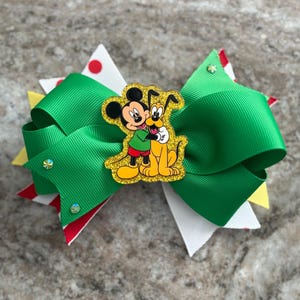May include: A green and white hair bow with a Mickey Mouse and Pluto embellishment. The bow features layers of green, white, yellow, and red ribbon, with a glittery Mickey Mouse and Pluto design in the center. Rhinestones add sparkle.