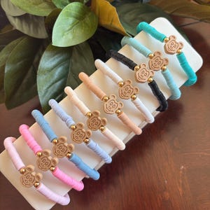 May include: A collection of colorful bracelets with a bear-shaped charm. The bracelets are in various pastel colors, including pink, blue, and white. Each bracelet has a gold-colored bead on either side of the bear charm. The bracelets are displayed on a white rectangular stand.