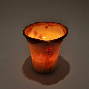 Könnte beinhalten: Ein hölzerner Kerzenhalter mit einem warmen, leuchtenden Inneren. Die natürliche Holzmaserung ist sichtbar, mit einer rauen, strukturierten Kante. Der Kerzenhalter wirft einen weichen Schatten auf die darunter liegende Oberfläche und erzeugt ein gemütliches Ambiente.