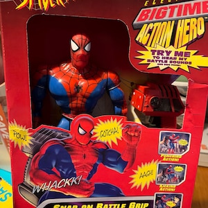Puede incluir: Un juguete Spider-Man Electronic Bigtime Action Hero en caja. El embalaje es rojo y presenta al personaje de Spider-Man con un traje rojo y azul. La caja incluye texto como "Try Me" y "Snap-on Battle Grip for Punching and Kicking Action!"