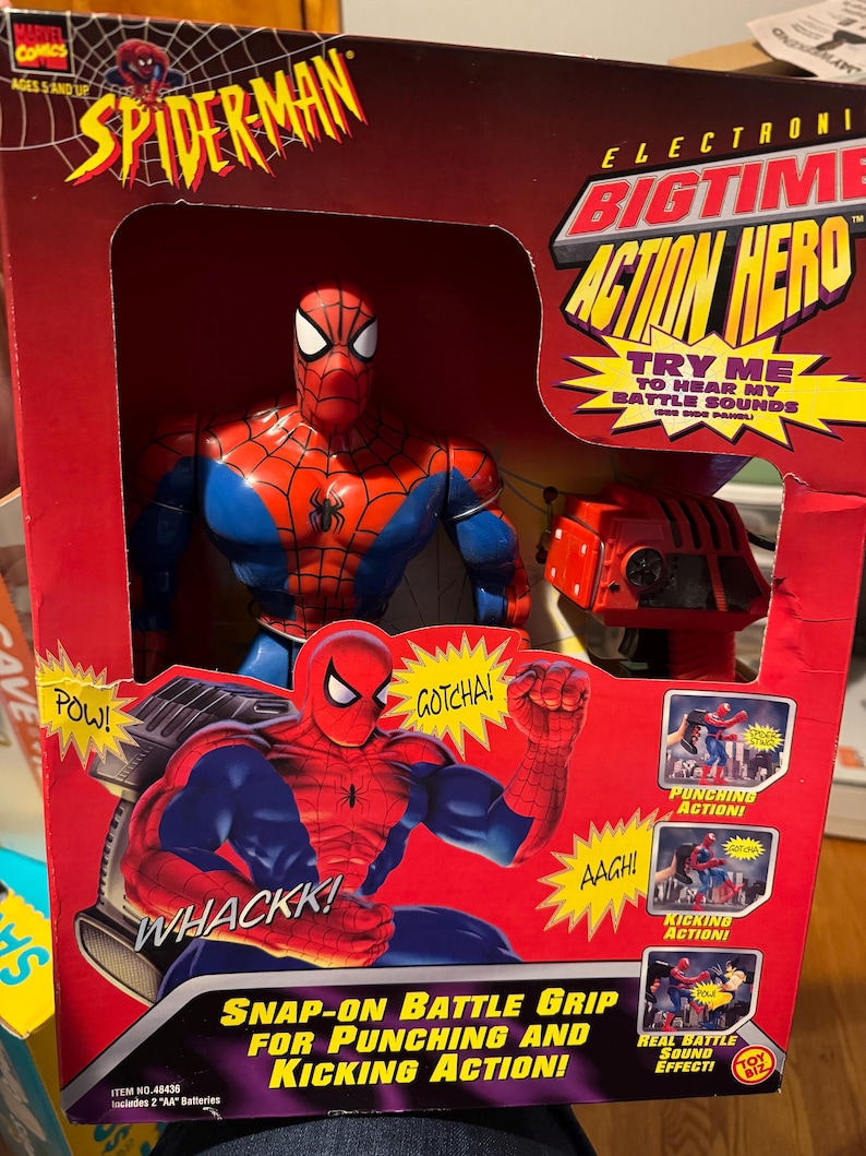 Puede incluir: Un juguete Spider-Man Electronic Bigtime Action Hero en caja. El embalaje es rojo y presenta al personaje de Spider-Man con un traje rojo y azul. La caja incluye texto como "Try Me" y "Snap-on Battle Grip for Punching and Kicking Action!"