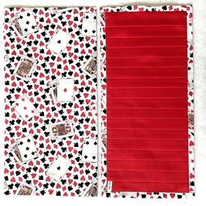 May include: A two-piece fabric item with a playing card pattern on one side and a solid red, pleated design on the other. The playing card side features a white background with red hearts, black spades, and playing cards.