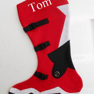 Peut inclure: Chaussette de Noël rouge avec des accents et boucles noirs, une bordure blanche et le nom "Tom" en blanc. La chaussette est conçue pour ressembler à une botte.