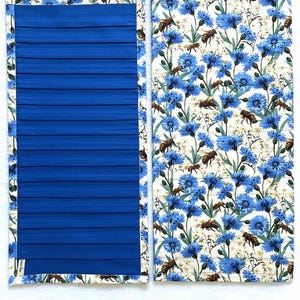 May include: Two rectangular fabric panels. One is solid royal blue with horizontal pleats. The other features a floral pattern of blue cornflowers, bees, and white accents on a cream background. Both have a patterned border.