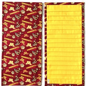 May include: A rectangular table runner featuring a maroon pattern with the word "MINNESOTA" and the letter "M" repeated. The reverse side is solid yellow with horizontal pleats. The edges are trimmed with the maroon pattern.