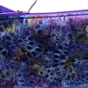 Könnte beinhalten: Ein rechteckiges Federmäppchen aus Stoff mit einem sich wiederholenden Muster aus violetten, blauen und türkisfarbenen abstrakten Blumenformen. Das Etui ist mit einem leuchtend violetten Reißverschluss an der Oberkante versehen, der leicht geöffnet ist.