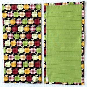 May include: Open notebook with a green lined paper interior. The cover features a black background with a pattern of red, green, and yellow gingham apples. The notebook is open, revealing the lined pages.