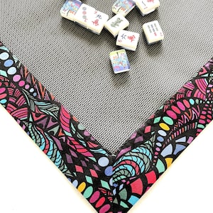 May include: A black mat with a colorful patterned border, perfect for playing mahjong. The mat has a grid pattern and is surrounded by a border with a variety of colors and shapes. There are several mahjong tiles scattered on the mat, showing Chinese characters and numbers.