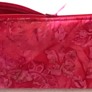 May include: A rectangular, zippered cosmetic bag in shades of pink and red. The bag features a floral pattern and a matching red zipper. The bag is designed to hold makeup and other beauty products.