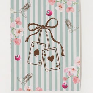 Puede incluir: Un cuaderno con un fondo a rayas azul claro y blanco, con ilustraciones de botas de vaquero, bolas de billar rosas y detalles florales. Dos cartas de juego, el As de Picas y el As de Corazones, están atadas con una cinta marrón.