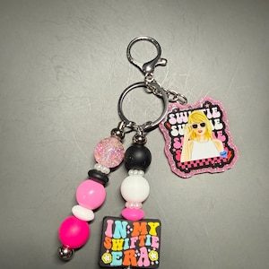 May include: A keychain with a silver clasp and ring, featuring a beaded strand with pink, white, black, and glitter beads. It includes two charms: one with a blonde woman in sunglasses and the text "Swiftie Summer," and another with the text "In My Swiftie Era."