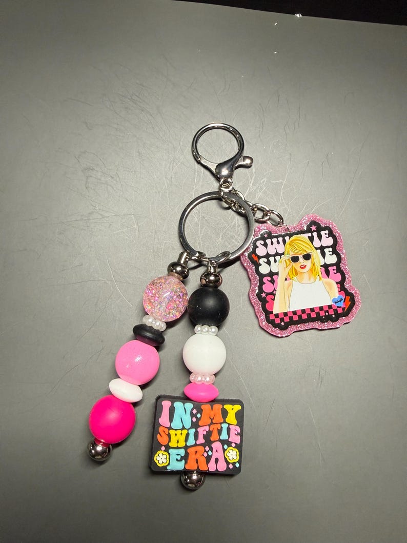 May include: A keychain with a silver clasp and ring, featuring a beaded strand with pink, white, black, and glitter beads. It includes two charms: one with a blonde woman in sunglasses and the text "Swiftie Summer," and another with the text "In My Swiftie Era."