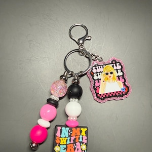 May include: A keychain with a silver clasp and ring. The keychain features a beaded strand with pink, white, black, and glittery beads. Attached are two charms: one with a cartoon image of a blonde woman and the words "Swiftie Summer", and another with the words "In My Swiftie Era".
