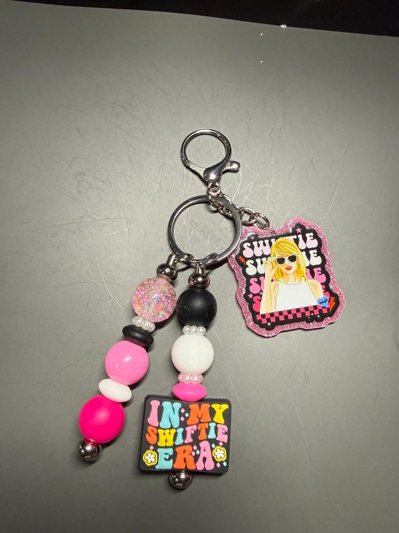 May include: A keychain with a silver clasp and ring. The keychain features a beaded strand with pink, white, black, and glittery beads. Attached are two charms: one with a cartoon image of a blonde woman and the words "Swiftie Summer", and another with the words "In My Swiftie Era".