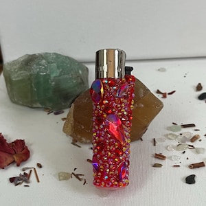 Könnte beinhalten: Ein rotes Feuerzeug ist mit roten und irisierenden Strasssteinen in verschiedenen Größen besetzt. Das Feuerzeug hat einen silbernen Metallaufsatz und liegt auf einem beigen Stein. Der Hintergrund umfasst einen grünen Stein und verstreute dekorative Elemente.