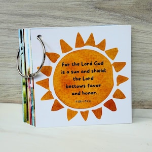 May include: A square card deck bound by a silver metal ring, featuring a watercolor-style orange sun illustration on the front cover. The text reads: For the Lord God is a sun and shield; the Lord bestows favor and honor. -Psalm 84:11.